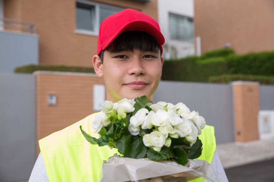 A young male flower delivery worker outdoors, wearing a red cap and a high-visibility yellow jacket, smiling while holding a bouquet of fresh white roses with green leaves wrapped in white paper. The