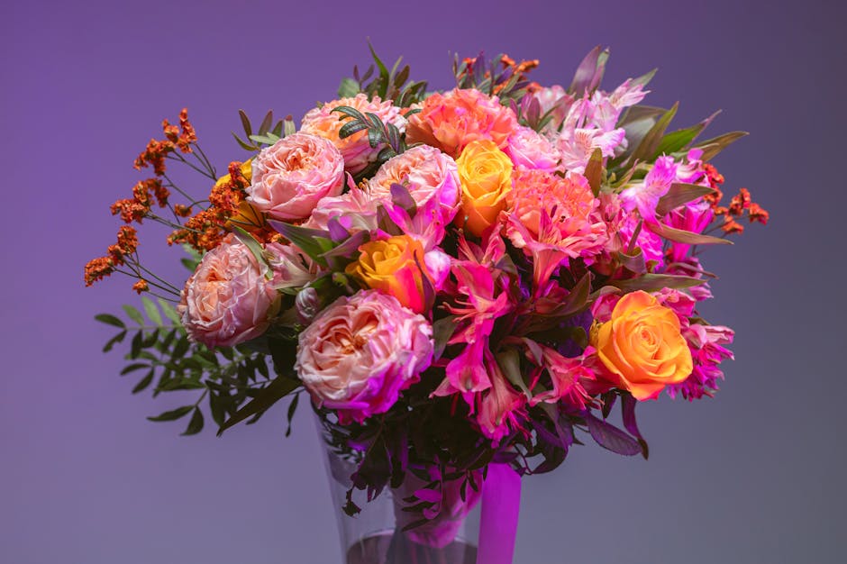 A vibrant floral arrangement featuring pink and peach roses, complemented by pink lilies and orange spray roses, all set within a clear glass vase. The bouquet is lush and full, with fresh, textured p
