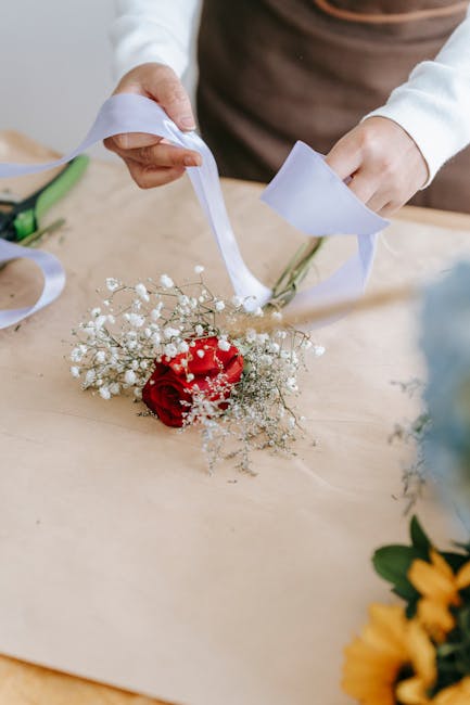 A person is arranging a floral gift on a wooden surface, holding a bouquet that features a vibrant red rose surrounded by delicate baby's breath and green foliage. The bouquet is wrapped with white ri