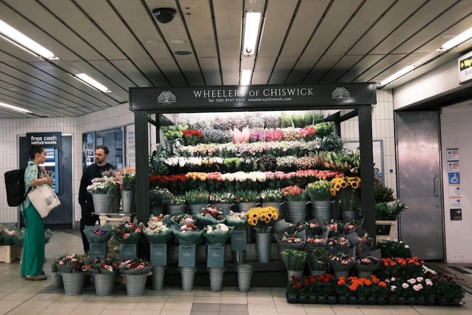 A floral display at Wheeler of Chiswick inside a subway station, presenting a diverse arrangement of fresh flowers including pink roses, white lilies, yellow sunflowers, orange marigolds, and purple t