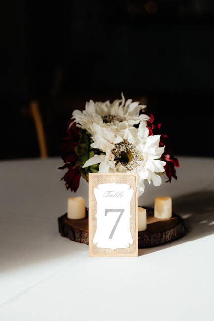 A floral arrangement serving as a table centerpiece featuring white daisies with dark centers and deep burgundy flowers, arranged in a compact bouquet. The flowers are set in a black vase or container